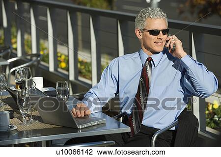 Stock Image - Prime adult Caucasian man in suit sitting at patio table ouside with laptop and talking on cellphone.. Fotosearch