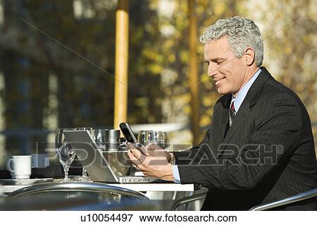 Stock Photo - Profile of smiling prime adult Caucasian man in suit sitting at patio table outside with laptop and dialing cellphone.. Fotosearch