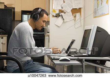 Stock Image - Side view of Asian young adult man sitting at desk typing on laptop wearing headphones.. Fotosearch