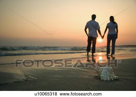 Stock Photo - Back view of mid-adult couple holding hands walking on beach with seashell in foreground.. Fotosearch