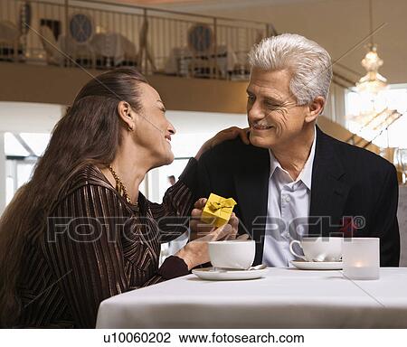 Stock Image - Caucasian mature adult male giving prime adult female gift box at restaurant table.. Fotosearch