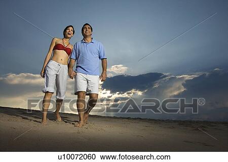 Stock Image - Caucasian mid-adult couple holding hands walking on beach.. Fotosearch