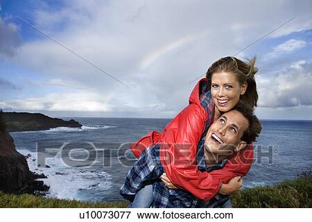 Stock Photo - Caucasian mid-adult couple piggybacking by ocean with rainbow in background in Maui, Hawaii.. Fotosearch