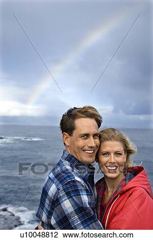 Stock Image - Caucasian mid-adult couple standing by ocean with rainbow in background in Maui, Hawaii.. Fotosearch