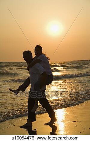 Stock Photograph - Caucasian mid-adult male carrying female piggyback on beach at sunset.. Fotosearch