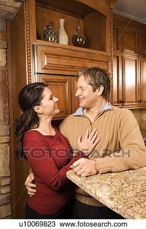 Caucasian woman and Caucasian man with arms around each other leaning on kitchen counter and smiling at each other. View Large Photo Image Stock Image - Caucasian woman and Caucasian man with arms around each other leaning on kitchen counter and smiling at each other.. Fotosearch