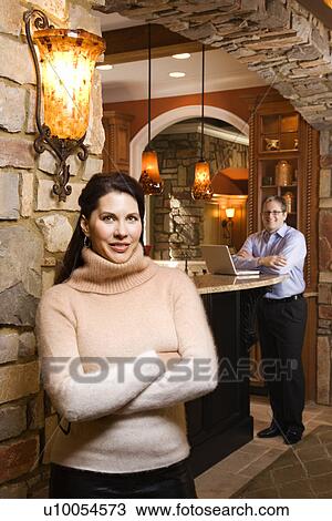 Stock Image - Caucasian woman standing with arms crossed looking at viewer with Caucasian man in background on laptop.. Fotosearch