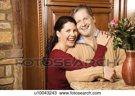 Stock Image - Caucasian woman with arms around Caucasian man in kitchen smiling and looking at viewer.. Fotosearch