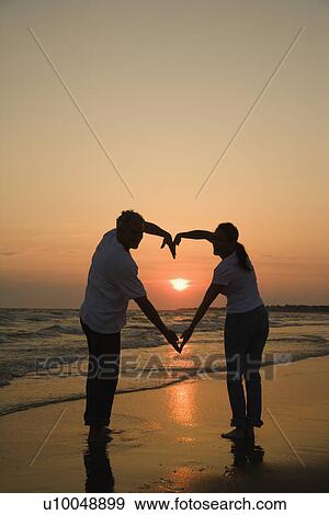 Stock Photo - Mid-adult couple making heart shape with arms on beach at sunset.. Fotosearch