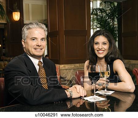 Stock Image - Prime adult Hispanic female and Caucasian prime adult male sitting at bar looking at viewer smiling.. Fotosearch