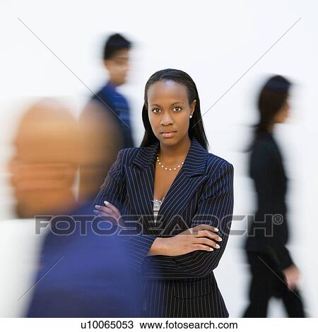 Stock Image - African-American businesswoman standing with arms crossed while others walk by.. Fotosearch