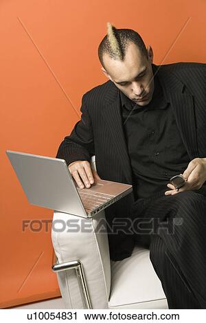 Stock Image - Caucasian man in suit with mohawk typing on laptop and looking at cellphone sitting in chair against orange background.. Fotosearch