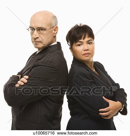 Stock Photograph - Caucasian middle-aged businessman and Filipino businesswoman standing back to back with arms crossed looking serious.. Fotosearch