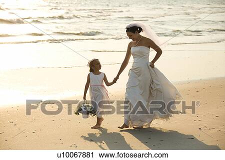 Stock Image - Caucasian mid-adult bride and flower girl holding hands walking barefoot on beach.. Fotosearch