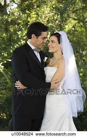 Picture - Caucasian mid-adult bride and groom embracing, smiling and looking at each other.. Fotosearch