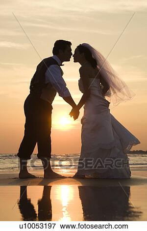 Stock Photo - Caucasian mid-adult bride and groom holding hands and kissing barefoot on beach at sunset.. Fotosearch