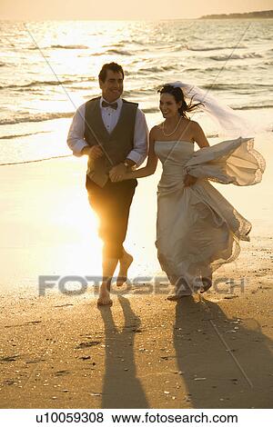 Caucasian mid-adult bride and mid-adult groom holding hands running barefoot on beach. View Large Photo Image Stock Photo - Caucasian mid-adult bride and mid-adult groom holding hands running barefoot on beach.. Fotosearch