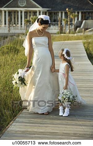 Stock Image - Caucasian mid-adult bride holding hands with flower girl walking down wooden beach walkway.. Fotosearch