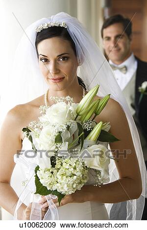 Stock Photo - Caucasian mid-adult bride holding bouquet with groom in background.. Fotosearch