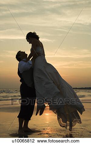Stock Image - Caucasian mid-adult groom lifting up bride on beach at sunset.. Fotosearch
