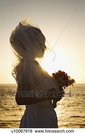 Stock Photo - Young adult female Caucasian bride at sunset holding bouquet on beach.. Fotosearch