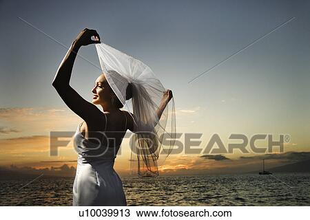 Stock Image - Young adult female Caucasian bride holding out veil on beach.. Fotosearch