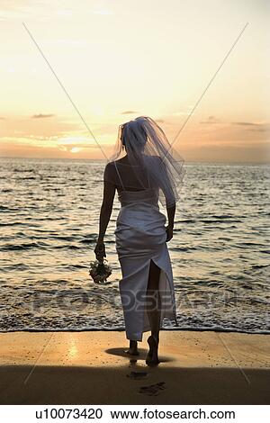 Stock Image - Young adult female Caucasian bride standing on beach at sunset.. Fotosearch