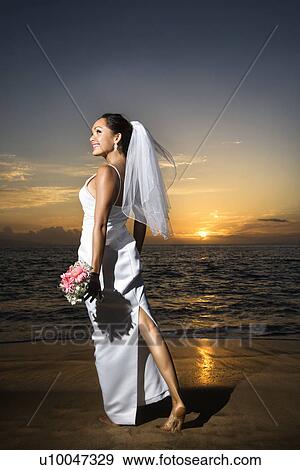 Stock Photo - Young adult female Caucasian bride standing holding bouquet on beach.. Fotosearch