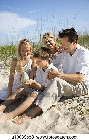 Stock Image - Caucasian family of four sitting on beach looking at seashell.. Fotosearch