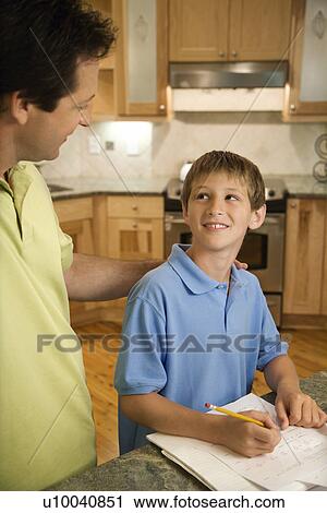 Stock Image - Caucasian mid-adult father helping pre-teen son with homework in kitchen.. Fotosearch