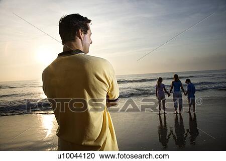 Stock Image - Caucasian mid-adult man standing and watching mid-adult woman with children on beach.. Fotosearch