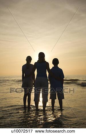 Stock Photo - Caucasian mid-adult mother and teenage kids standing silhouetted on beach at sunset.. Fotosearch