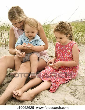 Caucasian mid-adult woman sitting with male toddler on lap and beside Caucasian female child on beach looking at shells. View Large Photo Image Stock Photo - Caucasian mid-adult woman sitting with male toddler on lap and beside Caucasian female child on beach looking at shells.. Fotosearch
