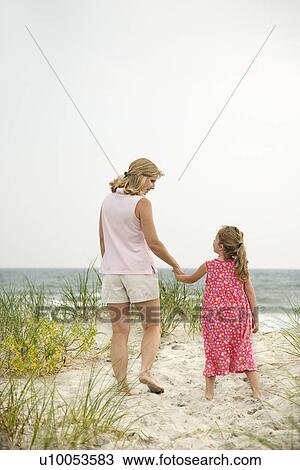 Stock Image - Caucasian mid-adult woman walking and holding hands with Caucasian female child on beach.. Fotosearch