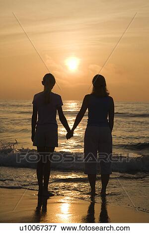 Stock Photo - Caucasian mid- female and teenage daughter standing on beach at sunset holding hands.. Fotosearch