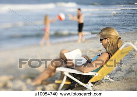 Caucasian mother reading in lounge chair on beach while husband and daughter play ball in background. View Large Photo Image Stock Image - Caucasian mother reading in lounge chair on beach while husband and daughter play ball in background.. Fotosearch