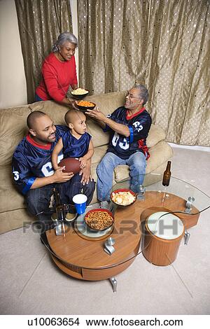 Picture - Mother serving snacks to male African-American family members during football game. . Fotosearch