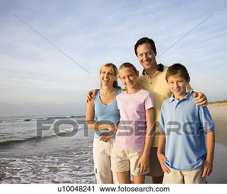 Stock Image - Portrait of Caucasian family of four posing on beach looking at viewer smiling.. Fotosearch