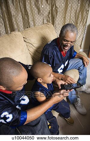 Stock Image - Three male generations of an African-American family watching football game on tv with boy holding remote.. Fotosearch
