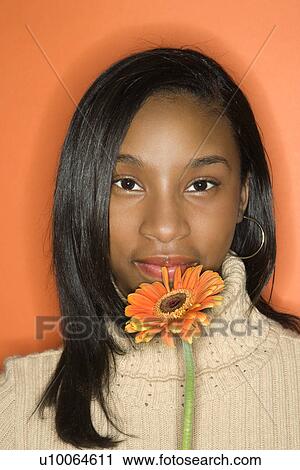 Stock Image - Portrait of African-American teen girl holding single Gerbera Daisy against orange background.. Fotosearch