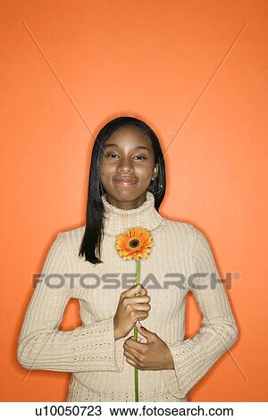 Stock Image - Portrait of grinning African-American teen girl holding single Gerbera Daisy against orange background.. Fotosearch