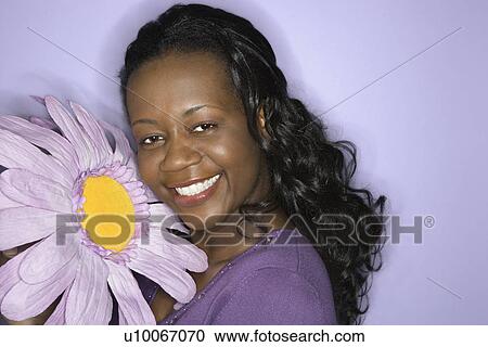 Stock Image - Portrait of smiling African-American mid-adult woman holding big purple fake flower on purple background.. Fotosearch