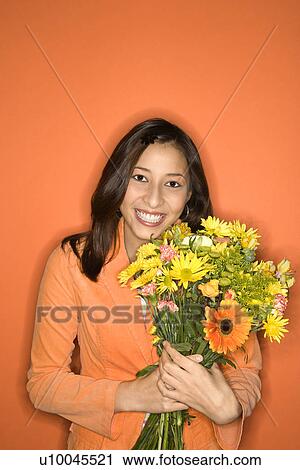 Stock Image - Portrait of smiling Multi-racial teen girl with bouquet of flowers standing against orange background.. Fotosearch