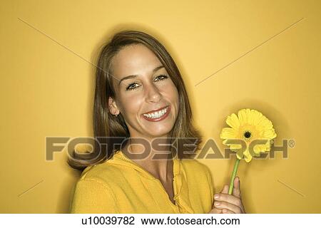 Stock Image - Portrait of smiling young adult Caucasian woman on yellow background holding flower.. Fotosearch