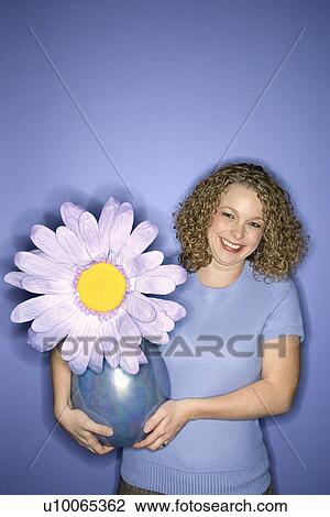Stock Image - Portrait of smiling young adult Caucasian woman on blue background holding flower pot with oversized fake flower.. Fotosearch