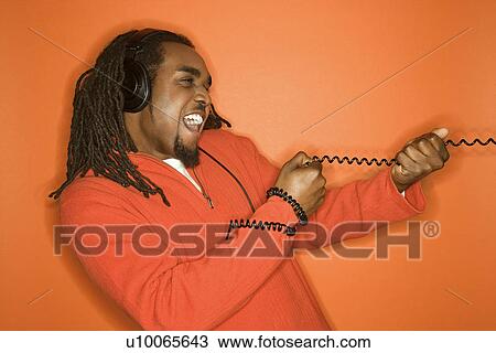 Stock Image - African-American mid-adult man pulling on cord wearing headphones and orange clothing on orange background.. Fotosearch