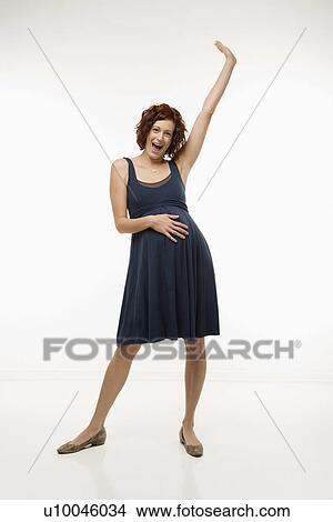Picture - Portrait of Caucasion mid-adult attractive pregnant woman standing with one hand on belly and other arm in air looking at viewer and smiling.. Fotosearch