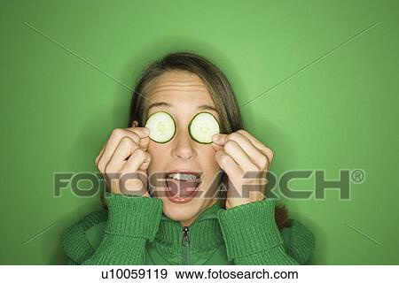 Stock Photo - Portrait of young adult Caucasian woman on green background holding cucumber slices over her eyes.. Fotosearch