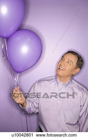 Stock Image - Smiling middle-aged Caucasian man on purple background holding purple balloons.. Fotosearch