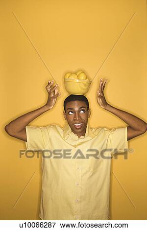 Stock Photo - Young African-American man balancing bowl of lemons on his head on yellow background.. Fotosearch